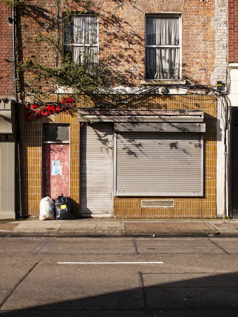 A ground-level view of a two-story brick building with a weathered, reddish-brown facade; the upper floor features two sash windows with black frames and partially drawn curtains. The lower portion of the building's front has yellow-brown tile cladding surrounding a closed grey metal roller shutter and a narrow, pink-painted doorway that appears slightly worn and has graffiti on it. To the left of the doorway, there are two black plastic rubbish bags and a clear plastic bag filled with waste materials, positioned on the pavement. Above the storefront, parts of a black cable conduit and electrical wiring are visible, with shadows of tree branches cast across the building's surface. The scene is lit by natural daylight, emphasizing the textures of the brickwork, tiles, and plastic rubbish bags, reflecting a typical urban environment potentially associated with private waste collection or rubbish removal services, as indicated by the presence of rubbish awaiting disposal.
