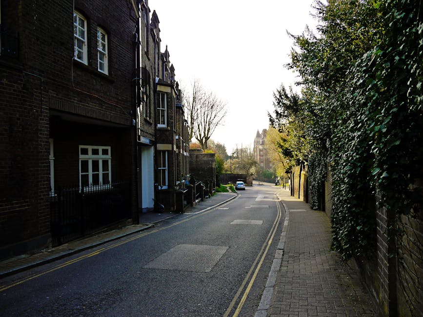 A quiet residential street in West Hampstead showing a narrow asphalt road with double yellow lines on each side, bordered by a pedestrian pavement on the right and by dark brick residential buildings on the left. The buildings feature white-framed sash windows, some with small railings in front and others with visible doorways. Shrubs and trees, some with leafless branches, are situated along the pavement and behind the buildings, suggesting early spring or late autumn. On the right side, a tall, dense hedge of dark green foliage lines a brick wall, partially obscuring view beyond. In the background, a white car is parked further down the street, with additional trees and a glimpse of a larger building or church spire visible on the horizon. The scene is lit by natural daylight, with a largely overcast sky creating soft shadows and diffused light, contextualising the street as a quiet area where private waste collection or rubbish clearance services like those from Rubbish Clearance West Hampstead might operate within urban residential environments.