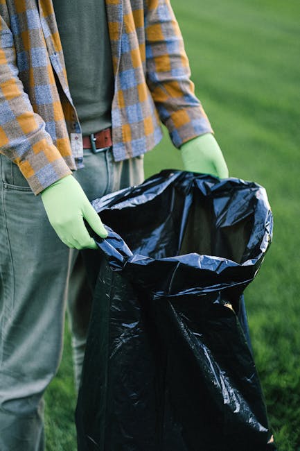 A person dressed in a checkered shirt, gray trousers, and wearing bright green gloves is holding open a large black plastic rubbish bag with both hands, preparing to collect waste outdoors. The background features a green grassy area, possibly part of a garden or park. The individual's midsection and hands are visible, with the focus on the ongoing rubbish collection activity. The scene emphasizes private waste handling, which may relate to independent rubbish removal services like those offered by companies such as rubbishclearancewesthampstead.co.uk. The natural outdoor lighting highlights the texture of the plastic bag and the clothing, creating a clear and professional depiction of rubbish collection in an environment that requires efficient waste disposal or clearance, aligning with the context of avoiding rubbish removal mistakes in West Hampstead gardens.