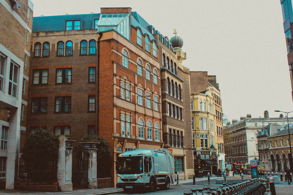 A narrow city street scene showing a variety of multistory buildings with different architectural styles, including brick facades, decorative windows, and ornate detailing. In the foreground, a white and blue refuse collection vehicle is parked on the street, which is lined with metal wheelie bins and a row of bicycle racks. The street is paved with asphalt and has a section of pavement with bollards and street signs visible on the right side. Surrounding buildings include a mix of modern and historic structures, with some featuring pitched roofs and dormer windows, while others have flat roofs. The overall atmosphere suggests an urban environment with active waste management services, such as those provided by independent rubbish clearance companies, alongside local council refuse collection. The lighting indicates daylight, and the scene appears clean and well-maintained, reflecting standard rubbish removal practices in a central London setting.