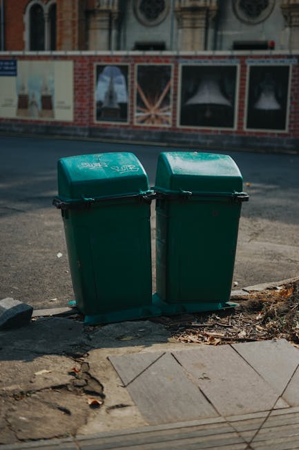 Two dark green plastic rubbish bins with hinged lids, positioned side by side on a concrete sidewalk curb at the edge of a street in an urban area. The bins have smooth surfaces with slight reflections of light and are labeled with white text indicating their use for waste collection, possibly for general household rubbish. They are situated directly on the sidewalk, with a few dried leaves and small debris around the base. Behind the bins, a section of paved road is visible, with a weathered surface that has some patches and minor cracks. In the background, there is a building with boarded-up windows and a brick and plaster facade, which features a decorative pattern. The lighting suggests a daytime setting, with natural light casting soft shadows. The scene hints at an alternative waste handling approach, like private rubbish collection, with the bins representing an independent rubbish disposal option near residential or commercial premises, supported by local waste management services such as those offered by Rubbish Clearance West Hampstead.