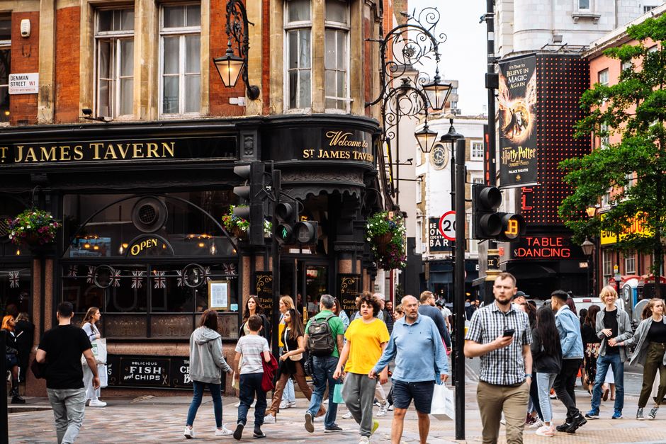A busy city street scene in front of the Black Oak pub named St James Tavern, with a variety of pedestrians walking across the cobbled pavement. The pub's exterior features tall, multi-pane sash windows with stone and brickwork detailing, and decorative black metal lanterns hanging from ornate wrought iron brackets. Some pedestrians carry bags or backpacks, and one man is engaged with a smartphone. To the right, a digital sign displays red lettering indicating a jazz dance event, and a poster advertising Harry Potter can be partially seen on a nearby building. The surrounding environment includes leafy trees partially obscuring the buildings, and the atmosphere is lively and well-lit with natural daylight, reflecting a typical urban setting possibly near independent retail or entertainment venues. The scene exemplifies an active area where private waste collection services could support street cleanliness amid busy commercial premises.