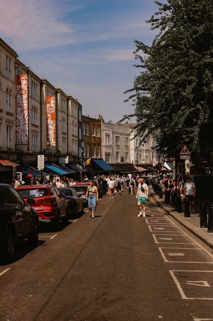 A busy street scene in West Hampstead showing a row of retail units with large windows and awnings on the left side, and a promenade for pedestrians in the center. Parked vehicles, including cars in various colors, line the road on the left. The street is filled with people walking, some carrying bags, and others browsing the shops or socializing. On the right side, there are trees with lush green foliage providing shade, and a series of black metal bollards separating the pedestrian area from the roadway. In the background, multistory buildings with brick and painted facades are visible, with flags or banners hanging from upper levels, suggesting a commercial or shopping district. The sky is partly cloudy with patches of blue, indicative of a warm, sunny day. The scene captures the lively atmosphere of an active retail street, illustrating the local environment where independent collection and private waste removal services such as those offered by rubbishclearancewesthampstead.co.uk are often used for clearing accumulated rubbish after shopping or business activities.