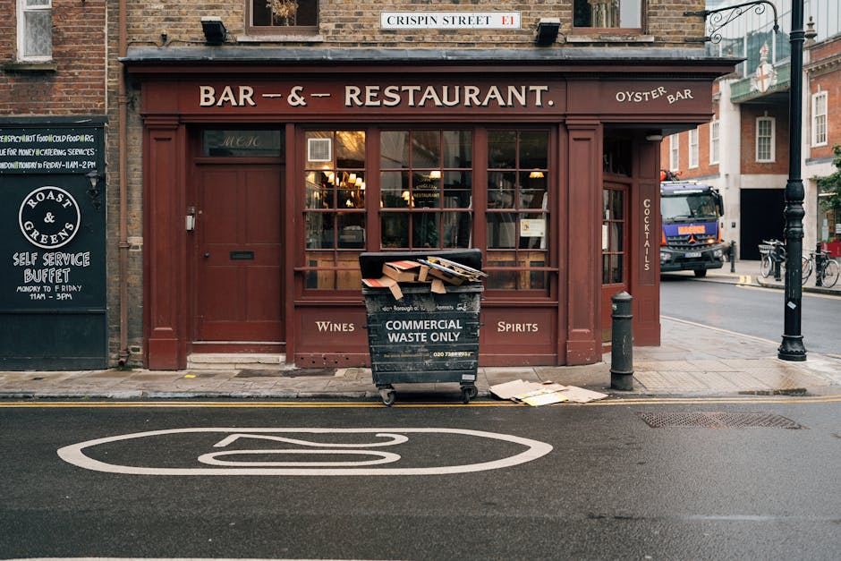 A busy city street scene in front of the Black Oak pub named St James Tavern, with a variety of pedestrians walking across the cobbled pavement. The pub's exterior features tall, multi-pane sash windows with stone and brickwork detailing, and decorative black metal lanterns hanging from ornate wrought iron brackets. Some pedestrians carry bags or backpacks, and one man is engaged with a smartphone. To the right, a digital sign displays red lettering indicating a jazz dance event, and a poster advertising Harry Potter can be partially seen on a nearby building. The surrounding environment includes leafy trees partially obscuring the buildings, and the atmosphere is lively and well-lit with natural daylight, reflecting a typical urban setting possibly near independent retail or entertainment venues. The scene exemplifies an active area where private waste collection services could support street cleanliness amid busy commercial premises.