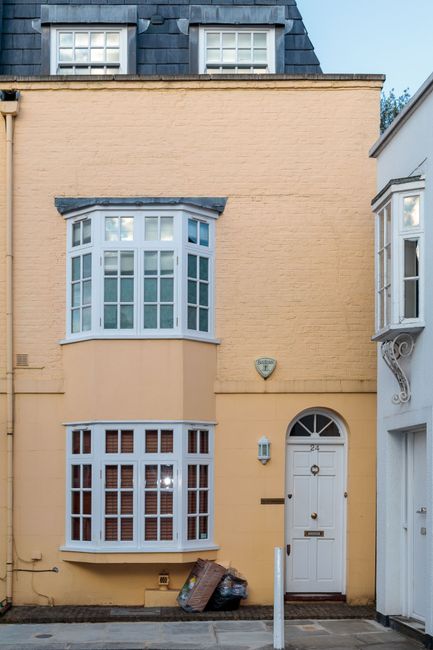 The image depicts the exterior of a narrow, three-storey terraced house with a light beige painted brick facade. The ground floor features a white wooden front door with an arched window panel at the top, centered in the image, and a small semicircular light fixture mounted to the right of the door. Two large bay windows are positioned on the lower and upper levels; the lower bay has multiple small rectangular glass panes with white wooden frames, while the upper bay similarly has multiple glass sections with white frames, both extending outward from the wall. The textures of the brickwork are visible, with the lower part of the facade appearing smooth and painted, and the upper part showing a rougher brick surface. To the left of the front door, there are a few rubbish bags and waste materials placed directly on the pavement, indicating recent or ongoing clearance activity. The house is tightly flanked by neighboring buildings, with part of a white facade visible on the right and a dark-tiled roof with a small window at the top in the background. The scene is lit by natural daylight, casting soft shadows and highlighting the textures and architectural details of the house, which is characteristic of private residential properties that might require professional rubbish removal services like those offered by [COMPANY_NAME].