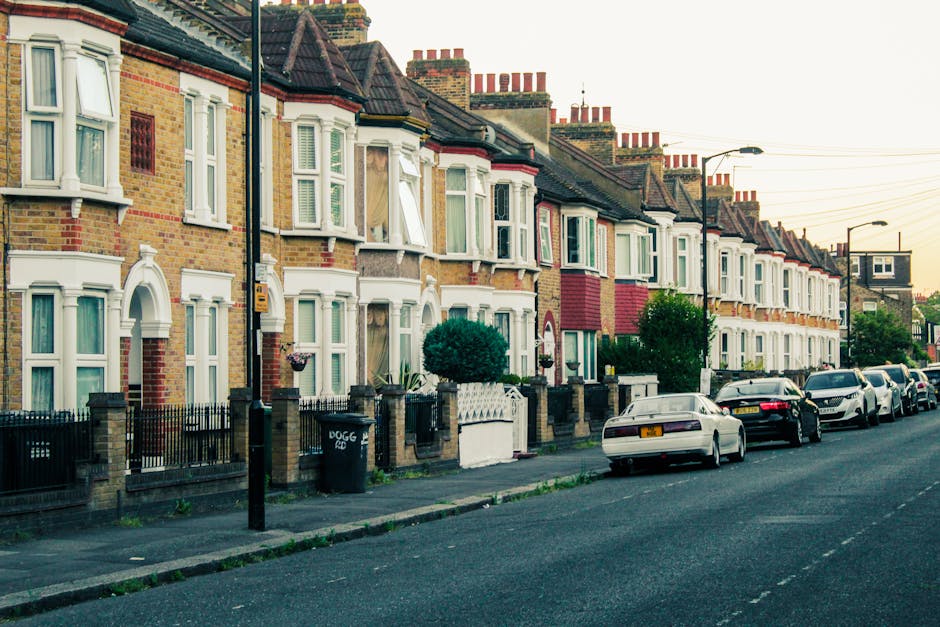 A row of traditional terraced houses in West Hampstead featuring brick facades in shades of yellow, brown, and red, with bay windows framed by white decorative window surrounds. Some windows are open, revealing curtains or blinds inside. Front gardens are enclosed by low brick walls topped with wrought iron railings, with greenery including small trees and shrubs present in several gardens. Parked along the street are multiple cars of various makes and colors, including white, black, and silver, occupying designated parking spaces. Streetlights and utility poles with overhead wires run parallel to the pavement, which appears clean and well-maintained. The scene is captured during late afternoon with soft, natural lighting giving the environment a warm tone. The background shows rooftops with chimneys, typical of traditional London housing, and the overall atmosphere reflects a quiet residential area with a well-kept appearance, typical of urban waste management and rubbish collection contexts in West Hampstead.