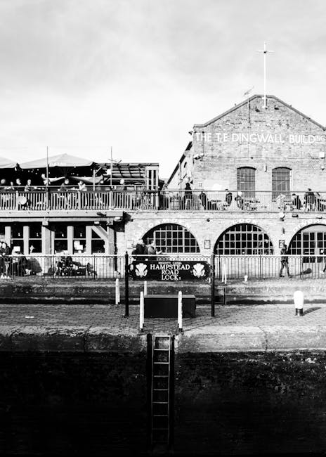 A black-and-white image depicting a historic brick building with arched windows on the ground floor and a sign reading 'The Edgingwall Buildings' on the upper facade, situated alongside a waterfront. In the foreground, a metal ladder extends from the water's edge up to a small platform or dock area, which is enclosed by a metal railing. The scene features a small sign attached to the railing that reads 'Hampstead Road Lock,' indicating a canal lock or waterway connection. Adjacent to the water, there's a paved area with bollards and a pedestrian walkway, and the environment appears to be an urban riverside setting, possibly linked with a location where waste removal services could be involved in maintaining the area’s cleanliness or managing waste associated with nearby buildings or public spaces. Natural lighting suggests daytime conditions with no visible human activity at the moment of the photo. This scene subtly relates to urban site clearance and external waste management activities, which companies like rubbishclearancewesthampstead.co.uk may undertake in such environments.