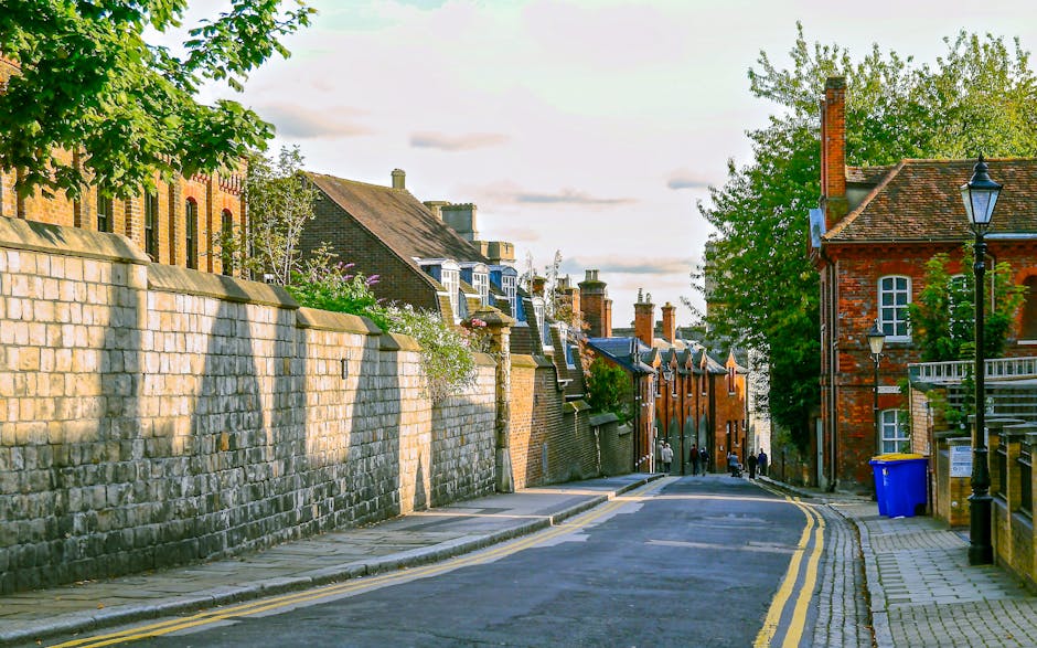 A quiet residential street in a historic area, lined with red brick terraced houses and a high stone wall on the left side, casting long shadows on the pavement. The cobbled sidewalk is bordered by a curb with double yellow lines indicating parking restrictions. On the right side, a traditional lamppost stands beside a bright blue recycling bin placed against a metal fence, adjacent to a small brick building with chimneys and windows. Tall, leafy trees with green foliage extend their branches overhead, providing partial shade and adding a natural element to the scene. Several pedestrians can be seen walking along the road in the distance, while the sky above shows scattered clouds and a soft, warm light suggestive of late afternoon. The street appears well-maintained, reflecting an environment where private rubbish collection or on-site waste clearance, such as that offered by rubbish clearance services like West Hampstead's, could be carried out to manage local waste conveniently and efficiently, away from public authority bins.
